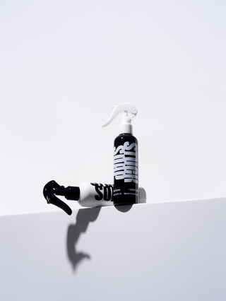 Black Silent Grove leave-in spray standing upright with a white Blush Garden spray lying on a ledge