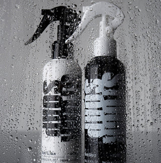 White Blush Garden and black Silent Grove leave-in spray bottles behind a pane covered in water droplets
